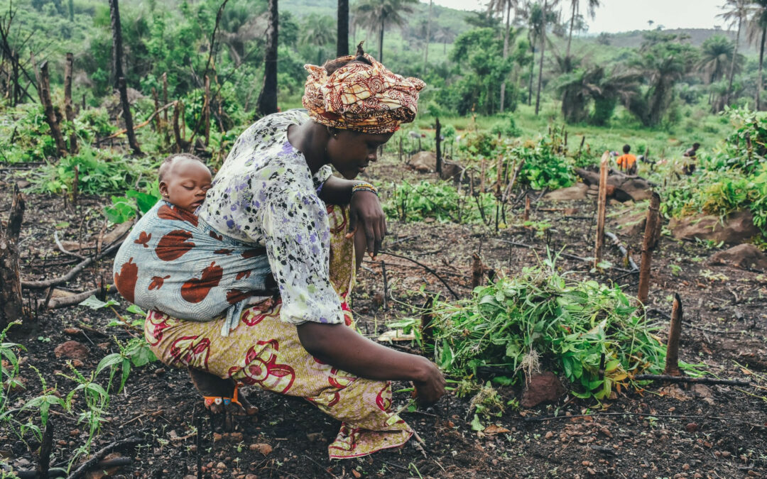 Donna che porta il bambino sulla sua schiena, con la tipica fasciatura africana, mentre coltiva il suo orto a mani nude.